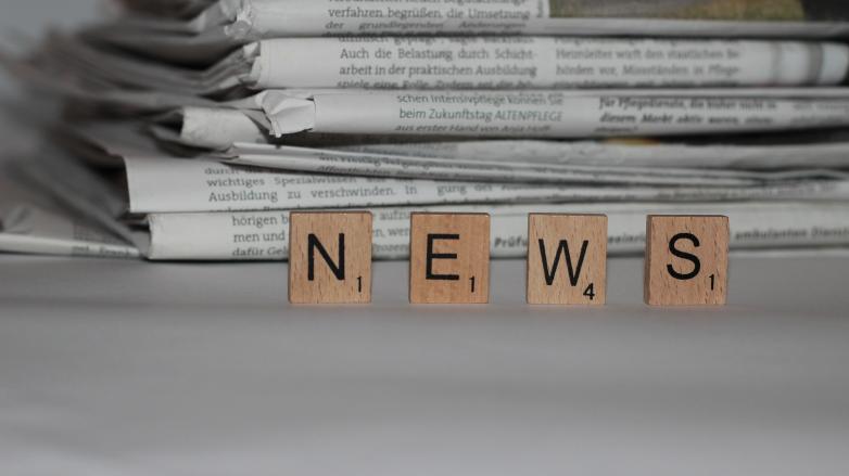 Das Wort "News" wird mit hölzernen Steinchen dargestellt, im Hintergrund sind Zeitungen zu sehen